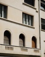 The image features a section of a building facade with a clean beige surface. Three arched windows, closed with wooden shutters, are evenly spaced on the lower level. Above them, a rectangular window is situated, composed of three vertical panes. Architectural details include a simple cornice and a vertical drainage pipe on the right.