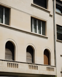 The image features a section of a building facade with a clean beige surface. Three arched windows, closed with wooden shutters, are evenly spaced on the lower level. Above them, a rectangular window is situated, composed of three vertical panes. Architectural details include a simple cornice and a vertical drainage pipe on the right.