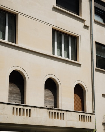 The image features a section of a building facade with a clean beige surface. Three arched windows, closed with wooden shutters, are evenly spaced on the lower level. Above them, a rectangular window is situated, composed of three vertical panes. Architectural details include a simple cornice and a vertical drainage pipe on the right.