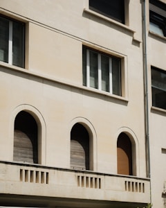 The image features a section of a building facade with a clean beige surface. Three arched windows, closed with wooden shutters, are evenly spaced on the lower level. Above them, a rectangular window is situated, composed of three vertical panes. Architectural details include a simple cornice and a vertical drainage pipe on the right.