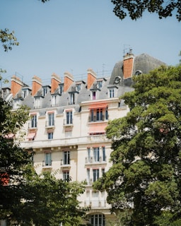 A European-style apartment building with intricate architectural details and red chimneys stands amidst lush green trees under a clear blue sky.
