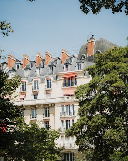 A European-style apartment building with intricate architectural details and red chimneys stands amidst lush green trees under a clear blue sky.