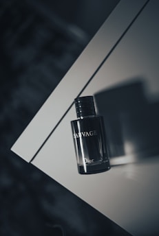 A bottle of Dior Sauvage perfume rests on a sleek, reflective surface with soft lighting creating a shadow on the table. The bottle is positioned at an angle, emphasizing its elegant design and dark color.