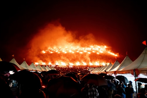 A large outdoor event with a significant display of flames on a hill in the background, causing an orange glow in the night sky. White tents and numerous umbrellas held by people can be seen in the foreground, suggesting it is raining.