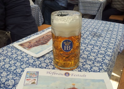 A large glass mug filled with beer, featuring a foamy head, sits on a table covered with a blue and white patterned tablecloth. The mug has a blue emblem with the letters 'HB' and 'Munchen' written on it. In front of the mug, there is a flyer or menu with the words 'Hofbräu Festzelt' printed on it. People are seated at the table, suggesting a festive or casual gathering.