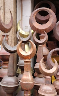 A collection of metal finials, predominantly featuring a crescent shape, displayed closely together. The items are made from different metals, showing a variety of tones from rusted brown to a brass-like gold. The setting suggests an outdoor market or a storage space.