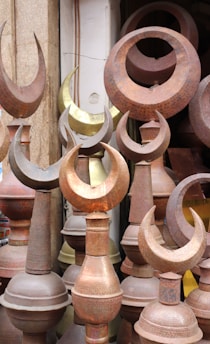 A collection of metal finials, predominantly featuring a crescent shape, displayed closely together. The items are made from different metals, showing a variety of tones from rusted brown to a brass-like gold. The setting suggests an outdoor market or a storage space.