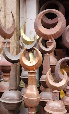 A collection of metal finials, predominantly featuring a crescent shape, displayed closely together. The items are made from different metals, showing a variety of tones from rusted brown to a brass-like gold. The setting suggests an outdoor market or a storage space.