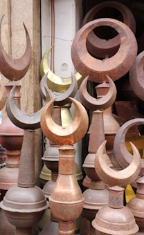 A collection of metal finials, predominantly featuring a crescent shape, displayed closely together. The items are made from different metals, showing a variety of tones from rusted brown to a brass-like gold. The setting suggests an outdoor market or a storage space.