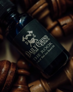 A close-up of a bottle of beard oil labeled 'Daily Grind Beard Co. The Rook' surrounded by wooden chess pieces. The lighting is moody, highlighting the metallic and matte texture of the bottle.