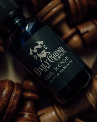 A close-up of a bottle of beard oil labeled 'Daily Grind Beard Co. The Rook' surrounded by wooden chess pieces. The lighting is moody, highlighting the metallic and matte texture of the bottle.