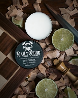 A tin of beard balm labeled 'Daily Grind Beard Co.' rests on a wooden surface surrounded by wood shavings and slices of lime. A wooden chess piece and a knife are also nearby.