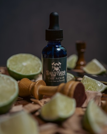 A bottle of beard oil labeled 'Daily Grind The Rook' is surrounded by several lime wedges and a wooden shaving brush. The bottle is a dark blue glass with a black dropper top. The setting is rustic, with wooden pieces scattered around.
