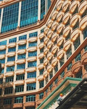 A striking building facade displaying a repetitive pattern of arched windows and geometric architectural features. The windows are accented with a peach-colored frame against a backdrop of light blue glass, reflecting the sky. In the bottom right corner, a person wearing a safety helmet and bright clothing is seen working on the building.