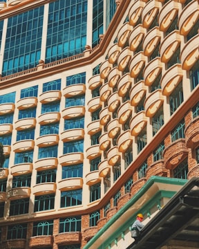 A striking building facade displaying a repetitive pattern of arched windows and geometric architectural features. The windows are accented with a peach-colored frame against a backdrop of light blue glass, reflecting the sky. In the bottom right corner, a person wearing a safety helmet and bright clothing is seen working on the building.