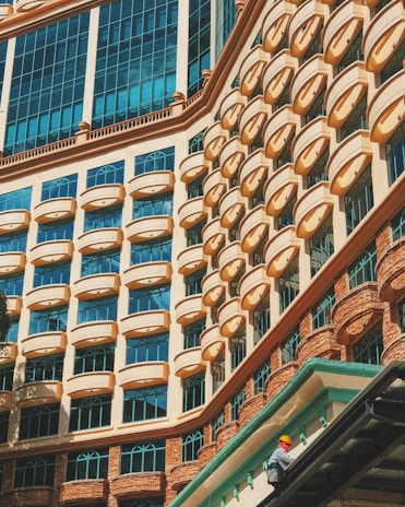 A striking building facade displaying a repetitive pattern of arched windows and geometric architectural features. The windows are accented with a peach-colored frame against a backdrop of light blue glass, reflecting the sky. In the bottom right corner, a person wearing a safety helmet and bright clothing is seen working on the building.