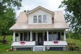 A two-story white house with beige metal roofing situated in a lush, green environment. The front features a covered porch with two large windows, two rocking chairs, and a welcome sign. American flag bunting decorates the porch railing, and the house number 4236 is prominently displayed on one of the support columns.