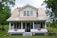 A two-story white house with beige metal roofing situated in a lush, green environment. The front features a covered porch with two large windows, two rocking chairs, and a welcome sign. American flag bunting decorates the porch railing, and the house number 4236 is prominently displayed on one of the support columns.