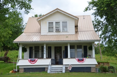 A two-story white house with beige metal roofing situated in a lush, green environment. The front features a covered porch with two large windows, two rocking chairs, and a welcome sign. American flag bunting decorates the porch railing, and the house number 4236 is prominently displayed on one of the support columns.