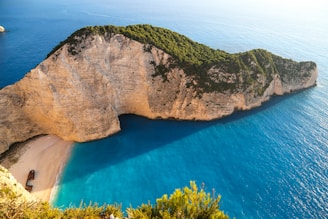 A breathtaking coastal landscape features a secluded beach bordered by steep, rocky cliffs covered with greenery. The pristine, turquoise sea waters create a striking contrast against the sandy shore and the cliff's rugged texture. A shipwreck rests prominently on the sand, adding an element of mystery to the serene environment.