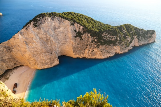 A breathtaking coastal landscape features a secluded beach bordered by steep, rocky cliffs covered with greenery. The pristine, turquoise sea waters create a striking contrast against the sandy shore and the cliff's rugged texture. A shipwreck rests prominently on the sand, adding an element of mystery to the serene environment.