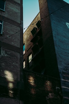 The image depicts an urban alley scene with tall, old brick buildings casting shadows. The lighting emphasizes the textures of the bricks, and there are dim lights strung across the gap between buildings.