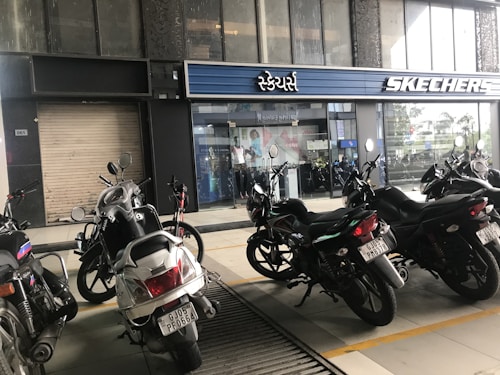 Several motorcycles are parked in front of a store with a sign reading 'Skechers' in English and another script. The storefront is modern with large glass windows and a roller shutter to the left. The area appears to be an urban environment.