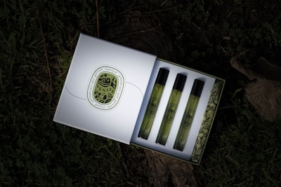 A box containing three perfume vials is open, revealing the vials neatly arranged in a row. Each vial has a greenish hue with black caps and is adorned with vintage-style labels. The box features an intricate design on the inside and a decorative emblem on the outside. It rests on a surface covered with grass and leaves, giving a natural, rustic backdrop.