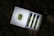 A box containing three perfume vials is open, revealing the vials neatly arranged in a row. Each vial has a greenish hue with black caps and is adorned with vintage-style labels. The box features an intricate design on the inside and a decorative emblem on the outside. It rests on a surface covered with grass and leaves, giving a natural, rustic backdrop.