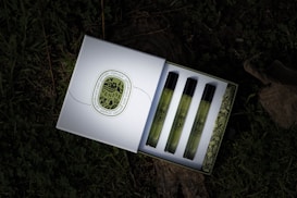 A box containing three perfume vials is open, revealing the vials neatly arranged in a row. Each vial has a greenish hue with black caps and is adorned with vintage-style labels. The box features an intricate design on the inside and a decorative emblem on the outside. It rests on a surface covered with grass and leaves, giving a natural, rustic backdrop.