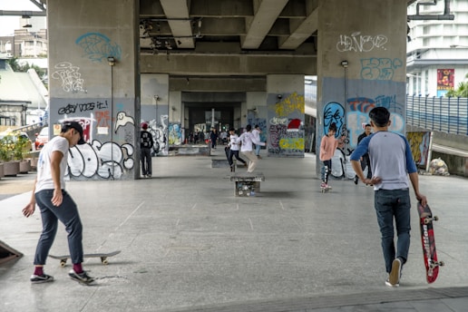 People are skateboarding under a bridge in an urban area adorned with graffiti. The scene is lively with individuals in casual attire engaging in skateboarding activities. A variety of graffiti art covers the concrete pillars and walls, adding a colorful backdrop to the setting.