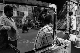 Several men are seen in an urban street scene, with one leaning against a window frame, another seated inside a vehicle, and others walking or standing nearby. A motorcycle is visible in the background, alongside a building and some plants.