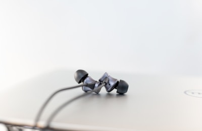 A pair of black wired earbuds are resting on top of a closed laptop with a plain, white background. The earbuds have a sleek, modern design with black rubber tips and a gray metallic finish. The laptop is slightly out of focus, revealing the corner of a logo.