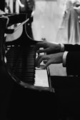 Hands playing a grand piano with an elegant and formal atmosphere, captured in black and white tones. The piano keys and hands are in sharp focus while the background remains softly blurred.