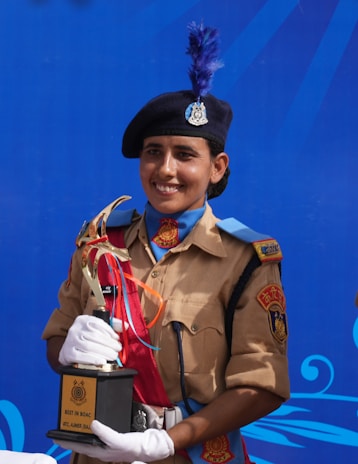A person in a uniform holding a trophy with a blue background. The uniform includes a beige shirt with decorative patches and a black beret with an insignia. They are wearing white gloves, and the trophy inscribed with 'Best in BOAC ATC Ajmer' features a gold top with ribbons.