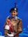 A person in a uniform holding a trophy with a blue background. The uniform includes a beige shirt with decorative patches and a black beret with an insignia. They are wearing white gloves, and the trophy inscribed with 'Best in BOAC ATC Ajmer' features a gold top with ribbons.