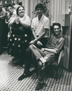 A group of people are gathered in a room with patterned flooring. One person is seated on a chair with their arm resting on the back, smiling. Another person stands beside them, wearing a white shirt. A third person is holding a drink and smiling, dressed in a polka-dotted outfit. The setting appears casual and social.
