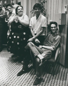 A group of people are gathered in a room with patterned flooring. One person is seated on a chair with their arm resting on the back, smiling. Another person stands beside them, wearing a white shirt. A third person is holding a drink and smiling, dressed in a polka-dotted outfit. The setting appears casual and social.