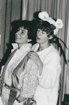 Two people are engaged in what appears to be a playful or theatrical interaction. One person is wearing a patterned robe and smiling while the other, dressed in a fluffy white outfit with bear-like ears, has their hands on the shoulders of the first person, with a slightly amused expression.
