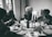 Four elderly individuals are seated around a dining table, enjoying a meal together. The table is set with various dishes, including matzah, and there are two lit candles in candlesticks in the center. The setting appears to be inside a home with windows showing an outdoor view.
