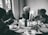 Four elderly individuals are seated around a dining table, enjoying a meal together. The table is set with various dishes, including matzah, and there are two lit candles in candlesticks in the center. The setting appears to be inside a home with windows showing an outdoor view.