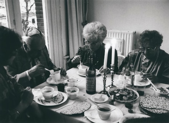 Four elderly individuals are seated around a dining table, enjoying a meal together. The table is set with various dishes, including matzah, and there are two lit candles in candlesticks in the center. The setting appears to be inside a home with windows showing an outdoor view.
