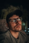 A man with a thoughtful expression is wearing glasses and a cap, captured in a dimly lit environment. Reflections of lights are visible in his glasses, adding depth to his introspective gaze.