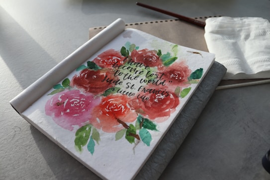 A notebook with a watercolor painting of roses on the open page is placed on a table. The painting includes various shades of red, pink, and green, with a quote written in cursive. Nearby, a paintbrush, a napkin, and a piece of textured paper are visible.
