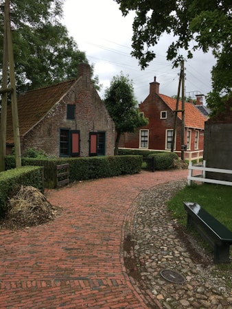 A narrow cobblestone path winds through a quaint village scene, passing traditional brick houses with steep roofs. Lush greenery surrounds the area, with trees and trimmed hedges lining the path. The atmosphere is serene and picturesque, evoking a sense of rural charm.
