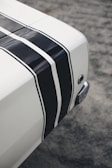 A close-up view of a part of a vehicle, featuring a white surface with bold black racing stripes running parallel across it. The texture of the ground beneath the vehicle appears rough and gray. There is also a metallic circular detail and a curved chrome element visible near the edge of the vehicle.