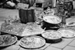 A street market stall features an array of handmade jewelry displayed on wooden platforms and circular trays. The crafts include necklaces and bracelets, emphasizing a rustic, natural aesthetic. Signs in the background suggest a theme of returning to nature. The setting has a casual, artisanal vibe.