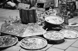 A street market stall features an array of handmade jewelry displayed on wooden platforms and circular trays. The crafts include necklaces and bracelets, emphasizing a rustic, natural aesthetic. Signs in the background suggest a theme of returning to nature. The setting has a casual, artisanal vibe.