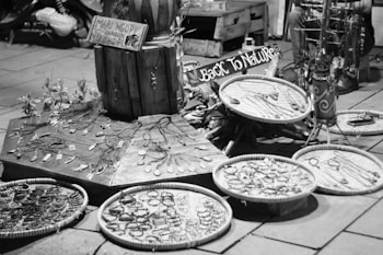 A street market stall features an array of handmade jewelry displayed on wooden platforms and circular trays. The crafts include necklaces and bracelets, emphasizing a rustic, natural aesthetic. Signs in the background suggest a theme of returning to nature. The setting has a casual, artisanal vibe.