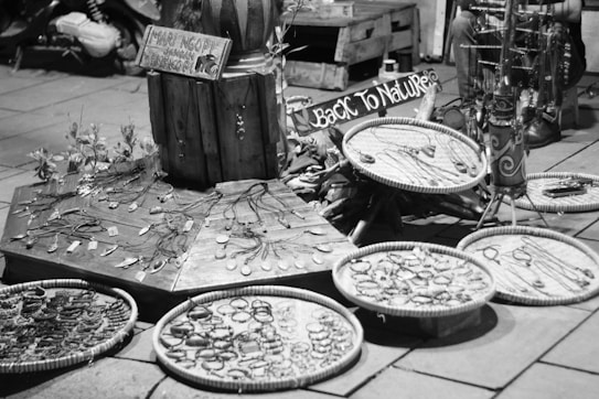 A street market stall features an array of handmade jewelry displayed on wooden platforms and circular trays. The crafts include necklaces and bracelets, emphasizing a rustic, natural aesthetic. Signs in the background suggest a theme of returning to nature. The setting has a casual, artisanal vibe.